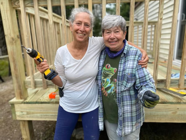 Two people stand with an arm over one another's shoulders and each hold a power tool. They smile at the camera with a completed porch behind them.