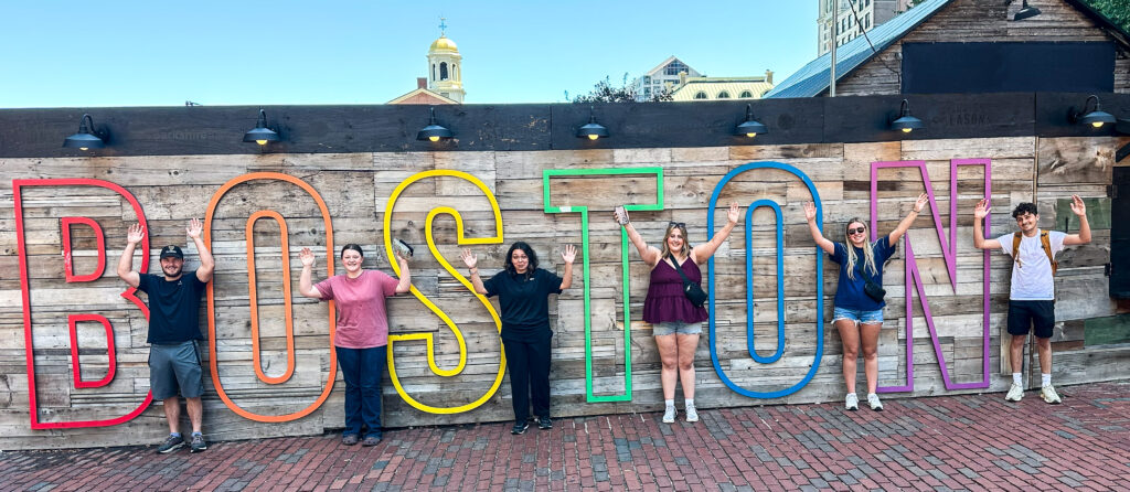 Six white older teenagers stand on a brick road in front of color lettering that spells out Boston.
